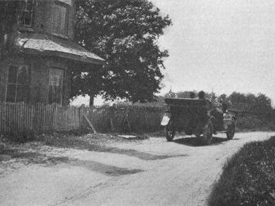 An early automobile travels on a rural road beside a building, fence, trees, and open land.