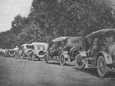 A line of early automobiles is parked along a rural road beside large trees.