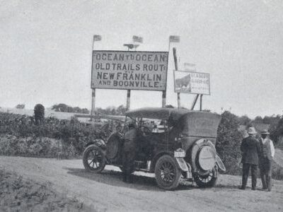 An early automobile is stopped beside 2 men near a roadside sign for the Old Trails Route through New Franklin and Boonville.