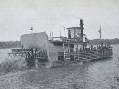 A paddlewheel ferry boat travels across a river with passengers on deck and water churning behind the wheel.
