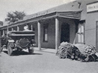 An early automobile is parked beside a long, covered building with a long covered porch, with a burro laden with several large bundles along the roadside.