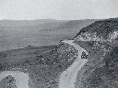 A narrow winding road curves along a steep hillside, with an early automobile traveling through an open mountainous landscape.