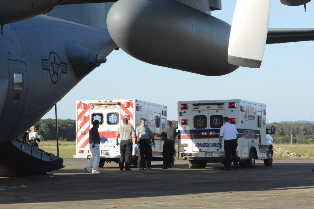 Two ambulances and several emergency personnel stand beside a large aircraft displaying a U S star-and-bar insignia on an airport apron.