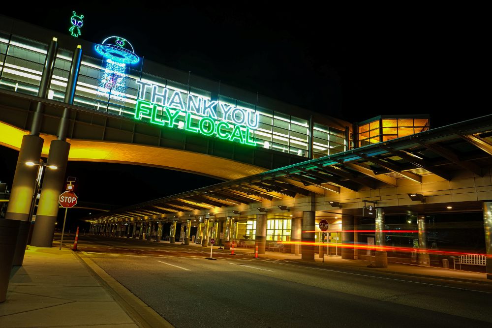 An airport terminal entrance at night has a large illuminated sign reading Thank You Fly Local above the roadway.