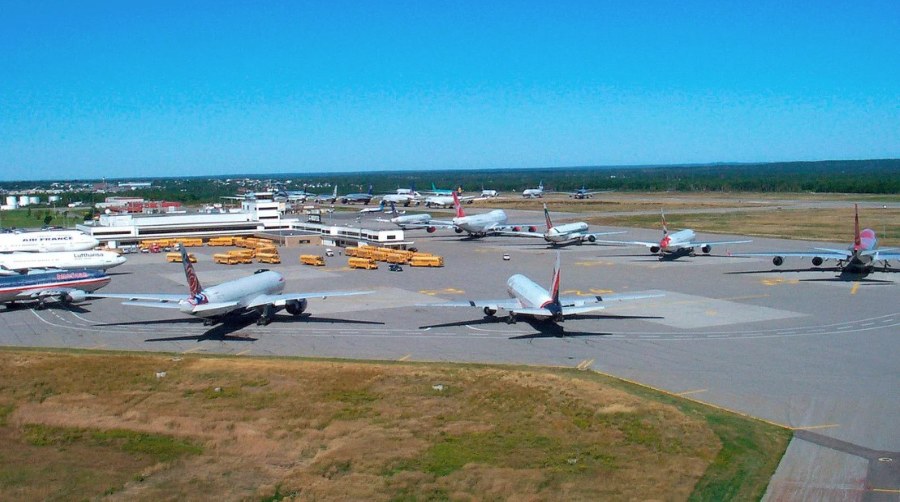 Several commercial passenger jets are parked wing-to-wing on an airport apron near terminal buildings, with school buses and other airport vehicles nearby.