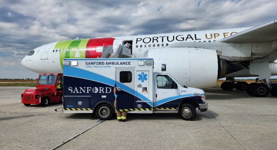 A Sanford ambulance stands beside a large TAP Portugal passenger airplane on an airport apron in Fargo, North Dakota, with a small service vehicle nearby.