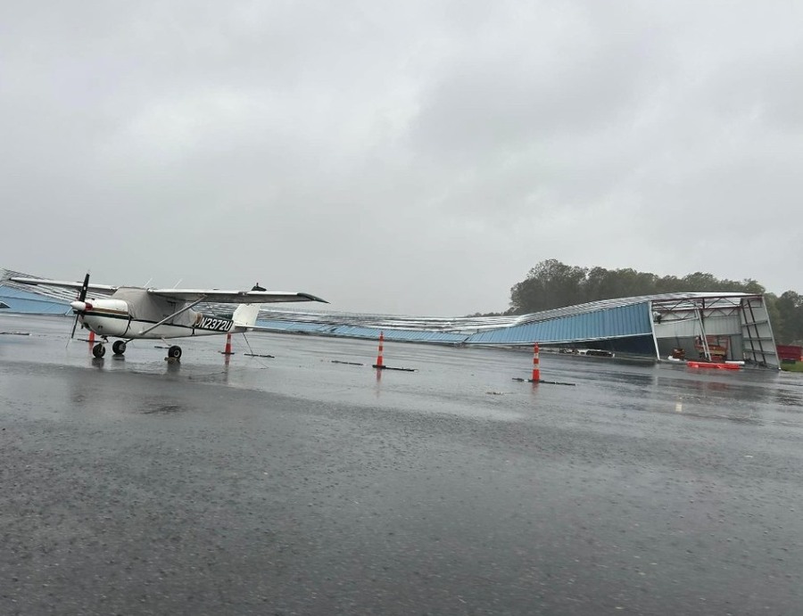 A small propeller airplane stands on a wet airport apron beside a partially collapsed hangar during rainy weather.