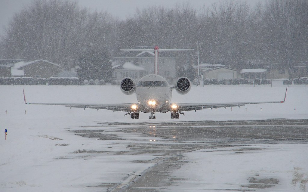 A small passenger jet taxis on a snow-covered runway during heavy snowfall with its landing lights on.