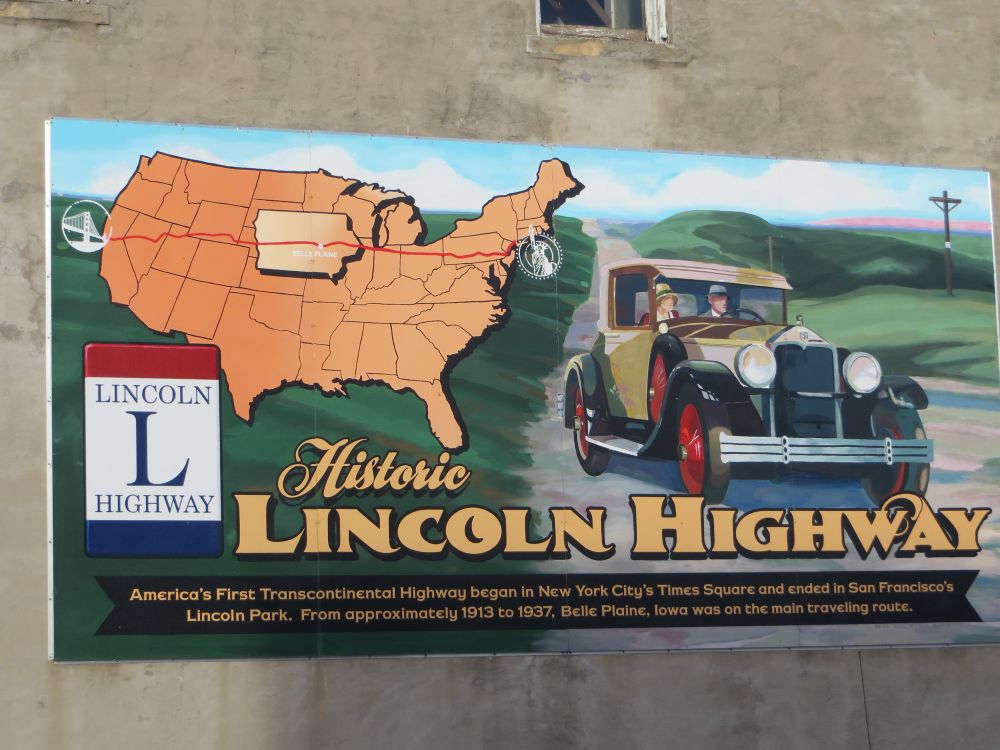 A Historic Lincoln Highway sign on the side of a building in Belle Plaine, Iowa, shows a U S map with the transcontinental highway route, a Lincoln Highway marker, and an early automobile.