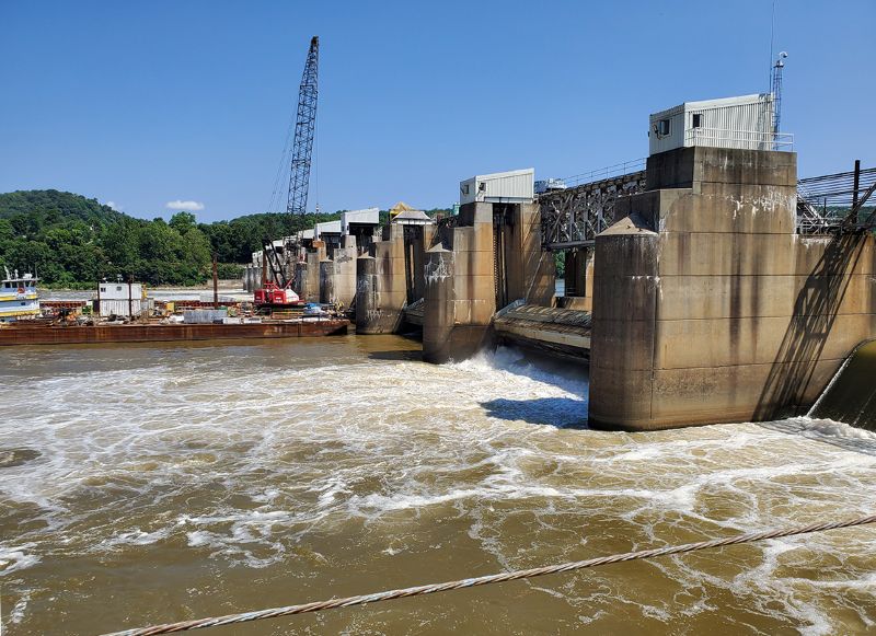 A dam structure spans a river with water flowing through openings beside large concrete supports, and a crane stands on a work area beside the dam.