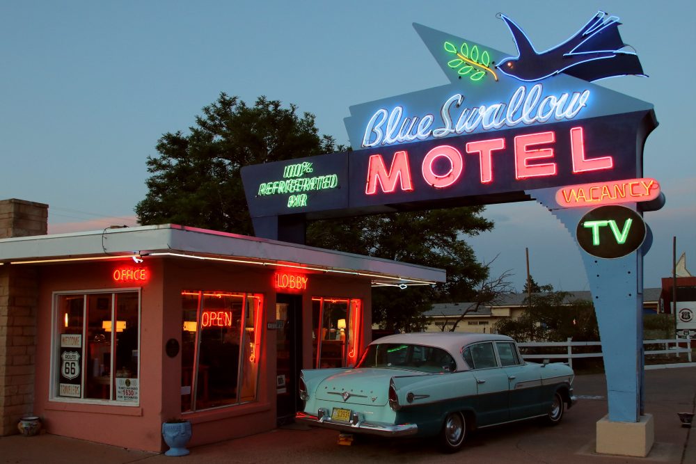 A Blue Swallow Motel sign advertises vacancy, T V, and 100 percent refrigerated air, with a vintage car parked outside the office.