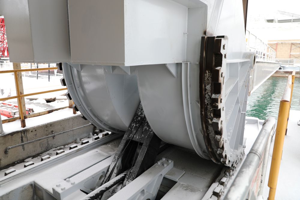 A large mechanical gate assembly sits on metal tracks beside water, with curved panels and support arms visible.