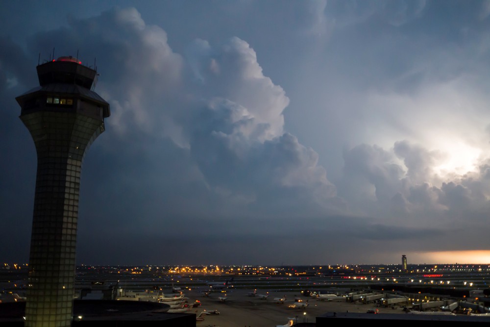 An airport control tower stands beside an airfield under large storm clouds, with aircraft and terminal areas visible below.