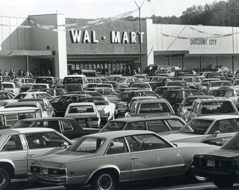 A crowded parking lot filled with 1980s vintage cars stands in front of a Wal-Mart store with Discount City signage.