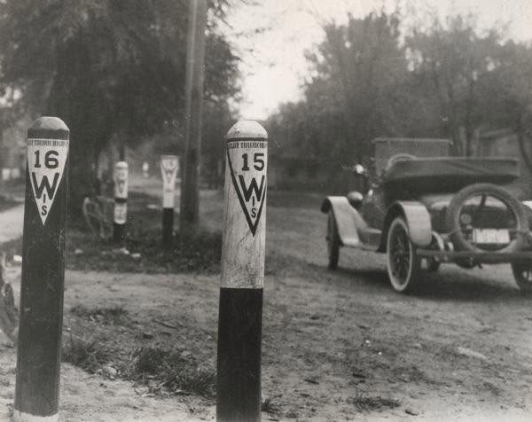 An early automobile travels on a rural road beside roadside markers labeled 15 W S and 16 W S.