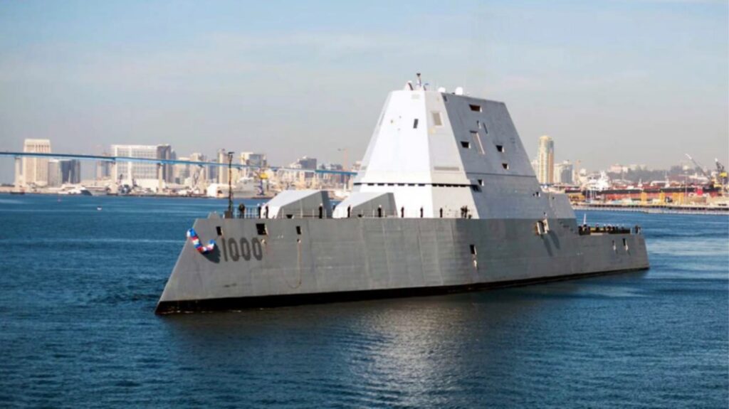 A large angular naval vessel marked 1000 moves through calm water with port buildings and structures in the background.