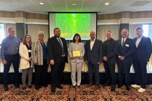 A group of nine people stands indoors as A C R P Manager Marci A. Greenberger at the center holds a 2025 Award of Excellence presented by the Michigan Aeronautics Commission and Michigan Department of Transportation.