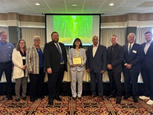 A group of nine people stands indoors as A C R P Manager Marci A. Greenberger at the center holds a 2025 Award of Excellence presented by the Michigan Aeronautics Commission and Michigan Department of Transportation.