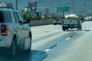 White pickup truck labeled 'WATER' sprays water onto the surface of a Utah highway with tiger-stripe lane markings. Behind that truck, another white utility vehicle equipped with a retroreflectometer gauges the reflectivity of the wet lane markings.