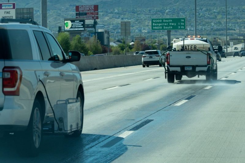 White pickup truck labeled 'WATER' sprays water onto the surface of a Utah highway with tiger-stripe lane markings. Behind that truck, another white utility vehicle equipped with a retroreflectometer gauges the reflectivity of the wet lane markings.