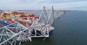 A large ship carrying stacked containers is under the Francis Scott Key Bridge collapsed over it in the water. Sections of the metal structure rest on the ship and extend into the water.