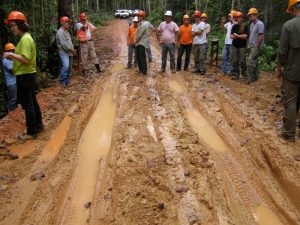 A muddy Amazon road is deeply rutted with water-filled tracks while a group of workers in helmets and safety gear stand observing the damaged surface.