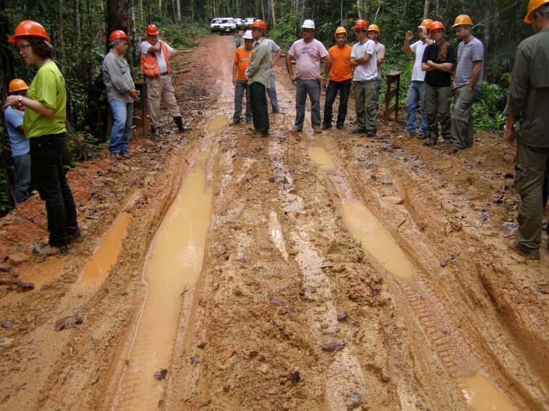 A muddy Amazon road is deeply rutted with water-filled tracks while a group of workers in helmets and safety gear stand observing the damaged surface.