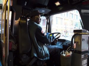 A bus driver wearing a cap and glasses sits at the wheel inside the driver’s cabin, steering through a city street visible through the front window.