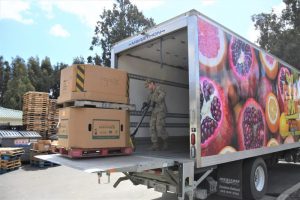 A person in a military uniform stands inside a truck and moves a pallet with stacked cardboard boxes of food using a hand pallet jack. Pallets and more boxes are in the background.