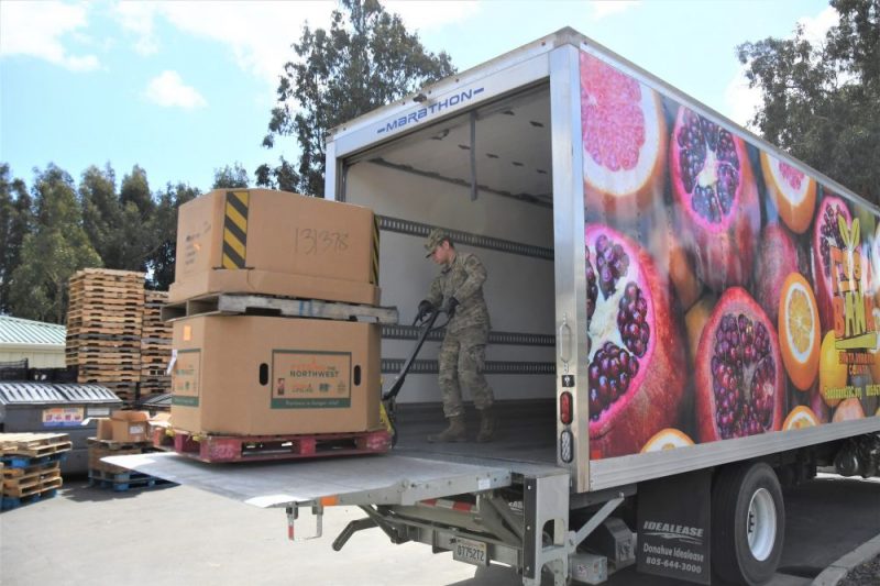 A person in a military uniform stands inside a truck and moves a pallet with stacked cardboard boxes of food using a hand pallet jack. Pallets and more boxes are in the background.
