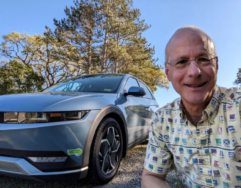 Doug Eisinger stands smiling beside a parked electric vehicle on a sunny day with trees in the background.