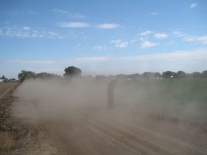 A person stands on a dusty unpaved low-volume road holding a tool while thick dust clouds rise, with fields and trees visible in the background.