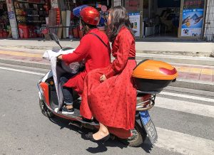 A person wearing a helmet drives an e-bike while another passenger in a red dress sits sideways on the back seat as they ride along a city street in China.