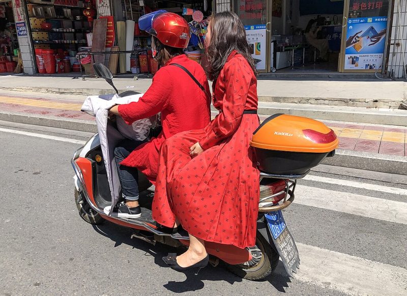 E-Bike Riders in Red A person wearing a helmet drives an e-bike while another passenger in a red dress sits sideways on the back seat as they ride along a city street in China.