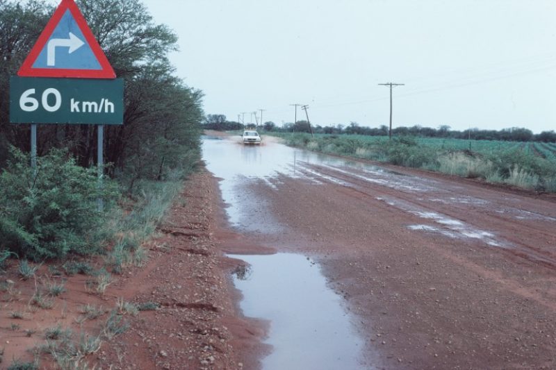 A car sprays water as it drives on a flooded, unpaved low-volume road in South Africa bordered by rows of crops and utility poles, with a speed-limit sign and scrub in the foreground.