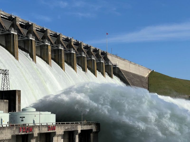 Water rushes down a dam spillway through several open gates and forms a large turbulent flow below.