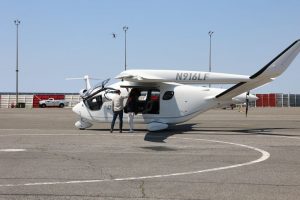 A small electric plane labeled N 916 L F sits on the tarmac at JFK International Airport with its door open while the pilot and a passenger stand beside the door.