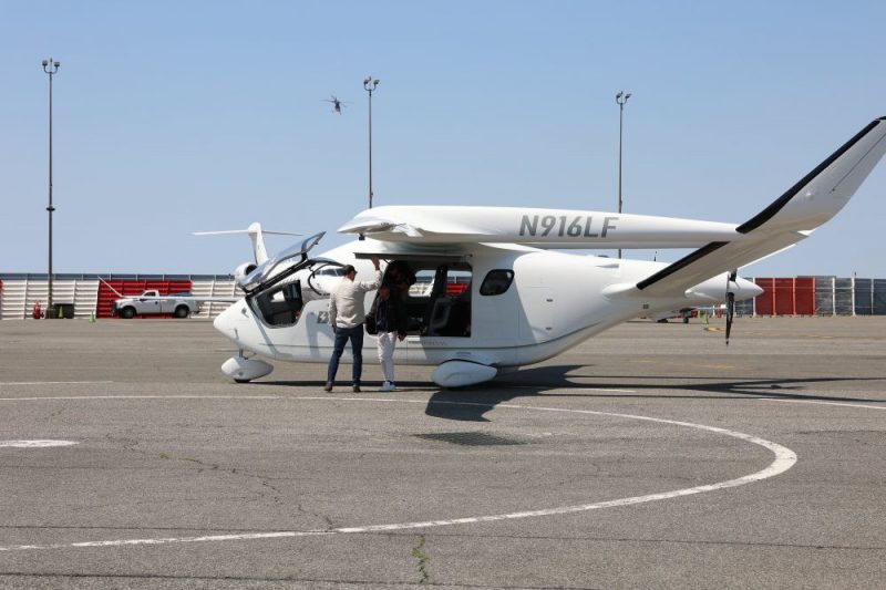 A small electric plane labeled N 916 L F sits on the tarmac at JFK International Airport with its door open while the pilot and a passenger stand beside the door.