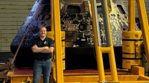 NASA engineer Jason Endsley stands with his arms crossed, smiling, in front of the cone-shaped crew module that carried the Artemis II astronauts around the Moon. The capsule sits on a hydrolic lift and has a patchwork of gold foil on tits charred exterior.