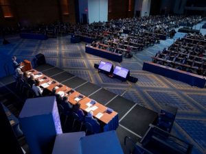 A large ballroom with many people seated at tables facing a stage where one speaker stands at the lectern and panelists sit at a long table.