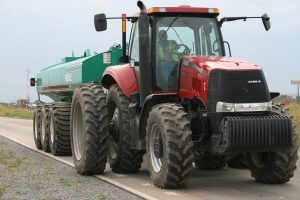 A large red tractor with large, dual rear wheels is shown pulling a green manure tanker along a paved road.