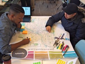 Two men sit across from each other while looking at a map of a draft bicycle network for the city of Minneapolis, Minnesota. The one wearing a blue cap and winter jacket points to a spot on the map while the other writes on an orage sticky note.