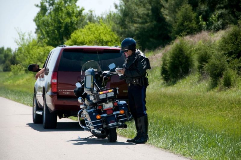 A Missouri state trooper stands near their motorcycle and writes on a notepad beside a stopped vehicle on a road.