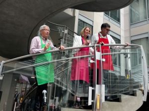 Three people wearing apron smocks stand on a spiral staircase. The man on the left holds a fudge bar. The woman in the center is speaking into a hand-held microphone. The man on the right has both hands on the railing and is looking downwards.