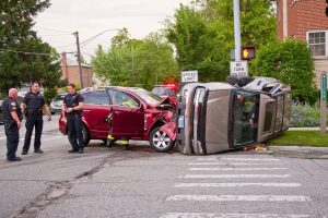 A car lies on its side with crushed roof and side, and another car with a smashed front end is T-boned into it while police officers stand around the intersection crash.