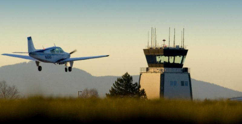 A small propeller plane descends over a grassy field toward the control tower at Hillsboro Airport near Portland, Oregon.