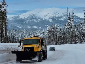 A snowplow sands a snowy mountain road with high snowbanks and snow-covered pine trees on either side. There is a car on the road behind the plow and a snow covered mountain topped by a wispy cloud.