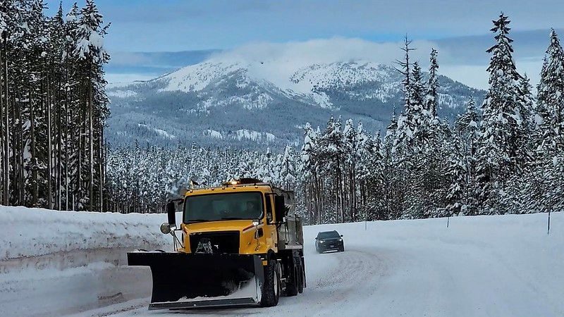 A snowplow sands a snowy mountain road with high snowbanks and snow-covered pine trees on either side. There is a car on the road behind the plow and a snow covered mountain topped by a wispy cloud.
