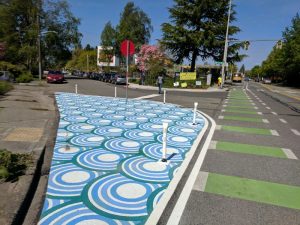 A street has a painted curb extension, bollards, tactile paving, and marked bike and crosswalk lanes.