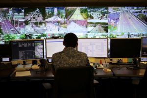 A person sits at a desk and looks at multiple screens. A large display wall shows many live road camera views.